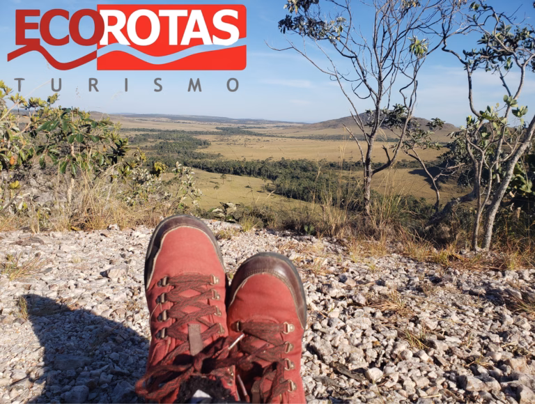 Par de botas de trilha vermelhas em primeiro plano. Elas estão voltadas para uma vista panorâmica de um vale do Cerrado na Chapada dos Veadeiros. No canto superior esquerdo, aparece o logotipo vermelho e branco da agência Ecorotas Turismo.