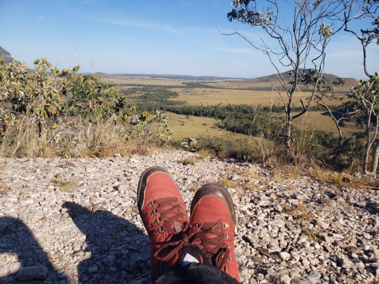 Fotografia em perspectiva de primeira pessoa mostrando as botas de trilha vermelhas de alguém sentado no topo do Morro da Baleia. Em primeiro plano, o chão é coberto de cascalho claro e pedras. Ao fundo, abre-se uma vista panorâmica de um vale plano e vasto, o Jardim de Maytrea, composto por vegetação rasteira de Cerrado em tons de dourado e verde, sob um céu azul claro e limpo logo após o amanhecer