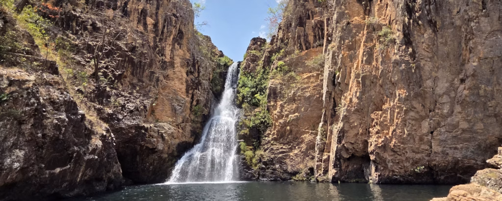 Vista frontal da Cachoeira do Encontro no Complexo Macaquinhos, na Chapada dos Veadeiros. A água flui em formato de véu branco entre imensos e altos paredões escarpados de rocha marrom. A queda deságua em um poço profundo e escuro sob um céu aberto com nuvens brancas. Foto por Ecorotas Turismo, Pousada Veadeiros e Norah Guia.