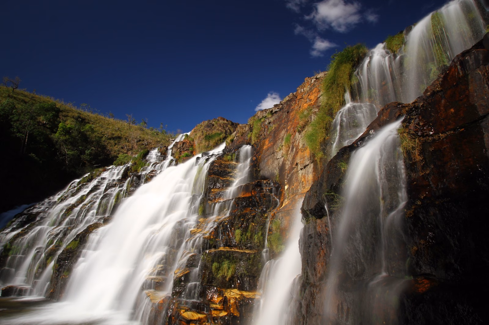 Cachoeira Almécegas Mil Catarata dos Couros I na Chapada dos Veadeiros com queda d'água em véu de noiva sobre paredão rochoso colorido, vegetação verde e céu azul intenso ao fundo.