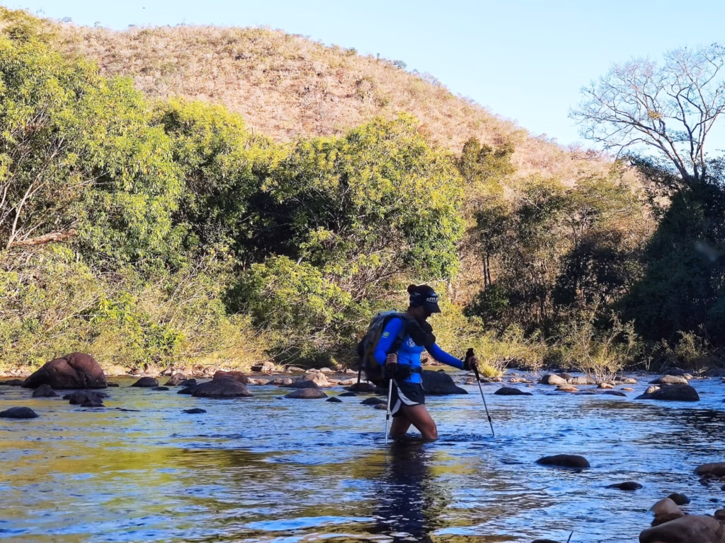 Pessoa faz travessia de rio a pé durante caminhada na natureza com morros ao fundo na Chapada dos Veadeiros